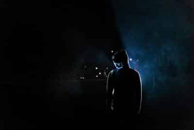 Silhouette of a man in a glowing LED mask and hoodie in a dark, foggy setting.
