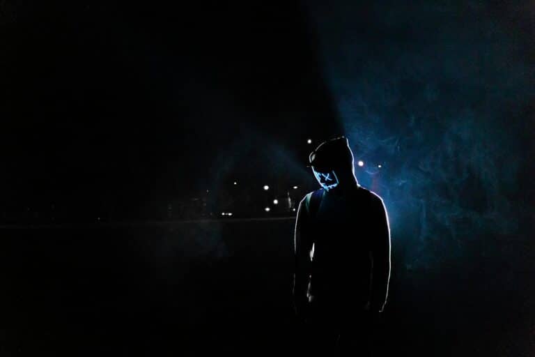 Silhouette of a man in a glowing LED mask and hoodie in a dark, foggy setting.