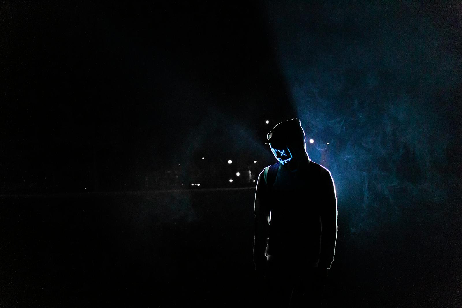 Silhouette of a man in a glowing LED mask and hoodie in a dark, foggy setting.